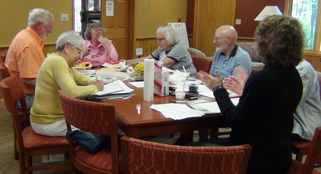 board-meeting-1024x555 A group of six older adults sitting around a table with papers and notebooks, in a warmly lit room.