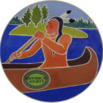 Native American male in a canoe with an orange outfit, paddling on a blue lake with green trees in the background.