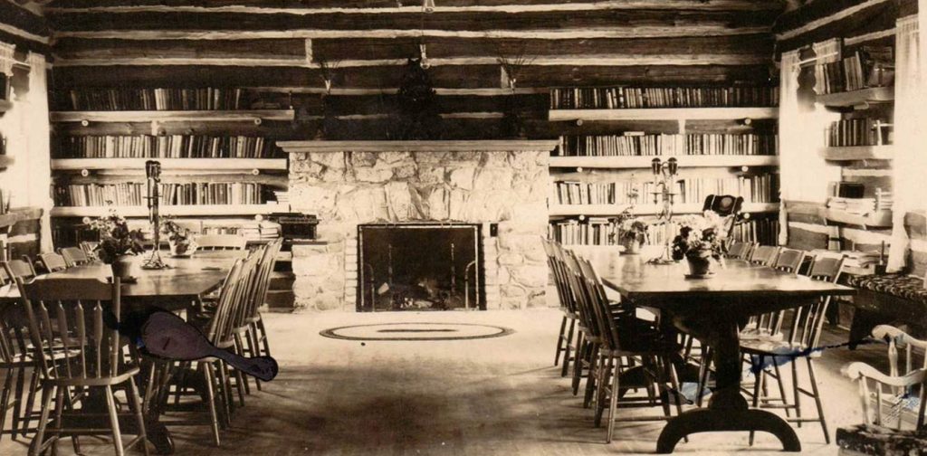 Rustic library in a log cabin with stone fireplace, wooden tables, and shelves filled with books.